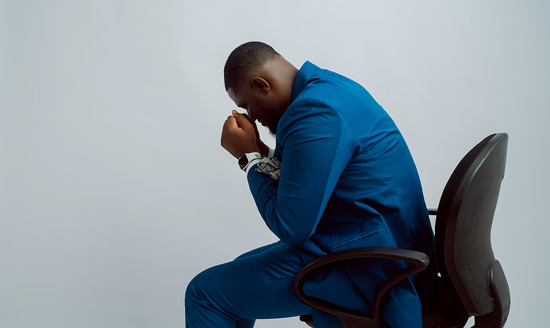 A person sitting hunched over a table, illustrating a prolonged bent back posture, with a subtle visual cue of strain on the lower and upper back muscles. The lighting highlights the stress points on the spine. Image must show ONLY what this section discusses: The connection between poor posture and upper and lower back pain, specifically prolonged sitting with a bent back, leading to muscle imbalance and increased pressure on the spine.. Photorealistic, high-quality photography style. אדם יושב כפוף ליד שולחן, מדגים יציבה לקויה היוצרת עומס על הגב העליון והתחתון.
